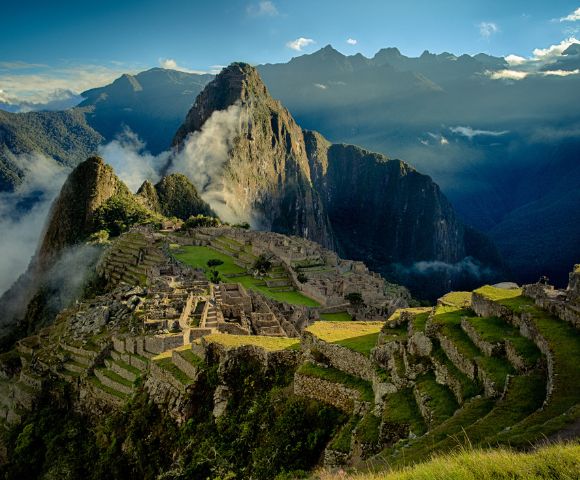 Alt text: A breathtaking view of Machu Picchu, with ancient stone ruins and terraces nestled in lush green mountains. The sky is partly cloudy, casting a serene, majestic glow over the landscape.