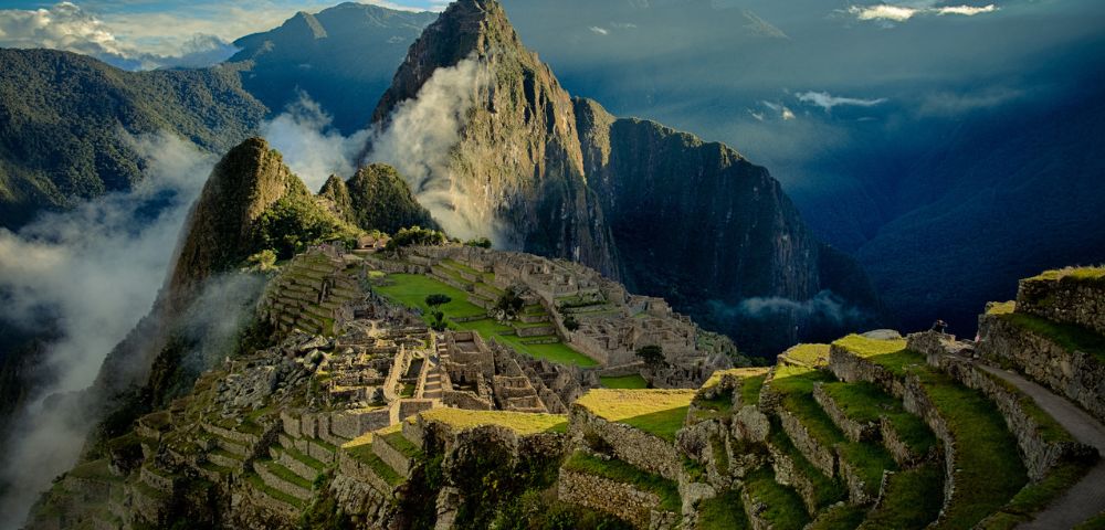Alt text: A breathtaking view of Machu Picchu, with ancient stone ruins and terraces nestled in lush green mountains. The sky is partly cloudy, casting a serene, majestic glow over the landscape.