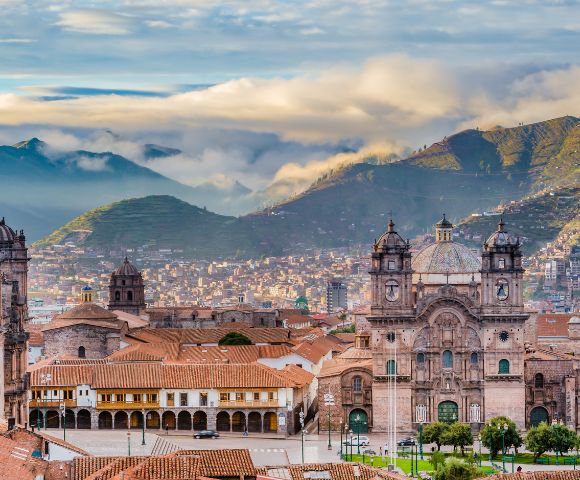 Historic cityscape of Cusco, Peru, with ornate cathedral towers and colonial buildings in the foreground. Misty mountains rise under a blue sky.