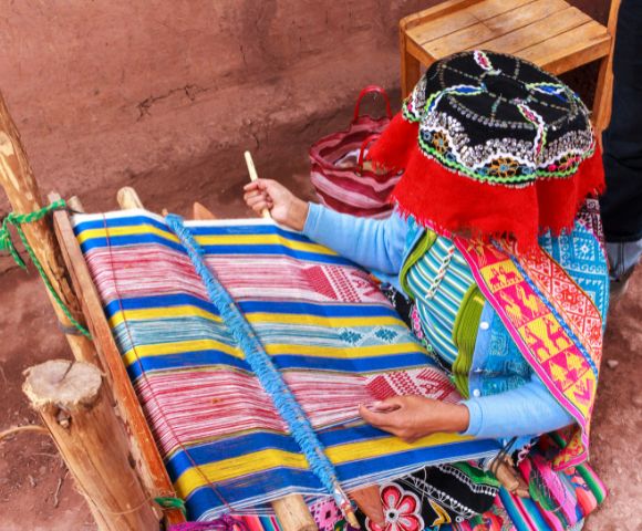 A woman in traditional attire weaves vibrant fabric on a wooden loom. She uses colorful threads, creating a detailed, geometric pattern. The scene is rich in culture and craftsmanship.