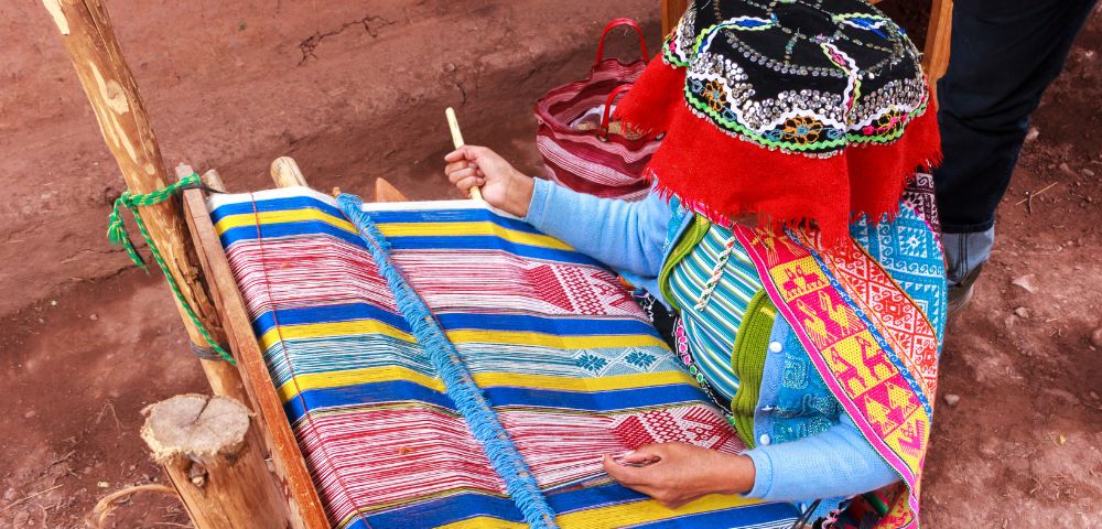 A woman in traditional attire weaves vibrant fabric on a wooden loom. She uses colorful threads, creating a detailed, geometric pattern. The scene is rich in culture and craftsmanship.