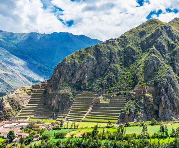 Terraced green mountainside with ancient ruins under a partly cloudy sky. Lush fields and small village at the mountain base. Historic landscape.