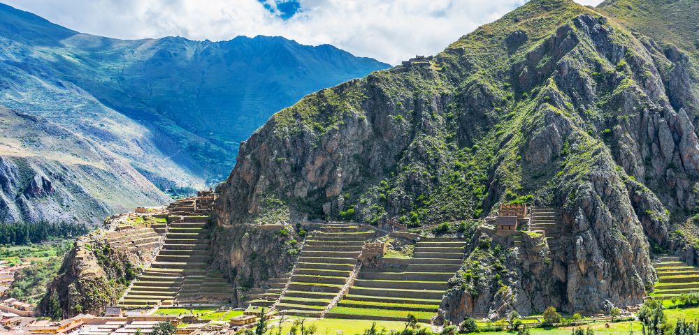 Terraced green mountainside with ancient ruins under a partly cloudy sky. Lush fields and small village at the mountain base. Historic landscape.
