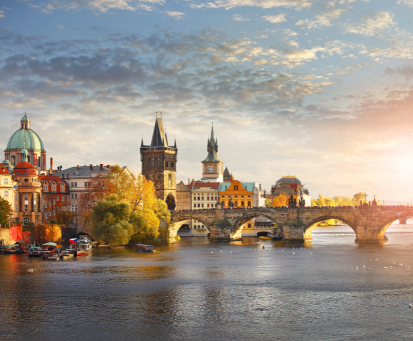 Panoramic view of Prague's Charles Bridge at sunset. Historic stone bridge flanked by Gothic towers, vibrant autumn trees, and tranquil river below.