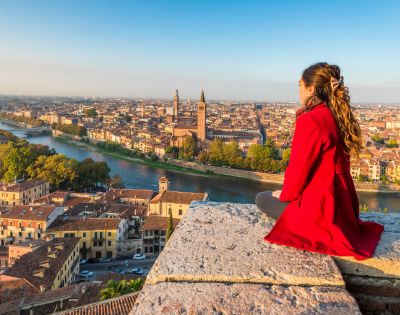 Young woman in a red coat sits on a stone ledge overlooking a scenic cityscape with a river, historic buildings, and distant towers under a clear blue sky.