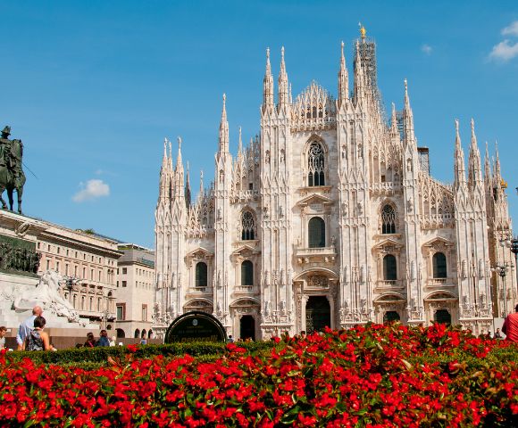 Gothic cathedral with intricate spires stands under a clear blue sky. Red flowers in the foreground and a statue to the left add contrast and depth.