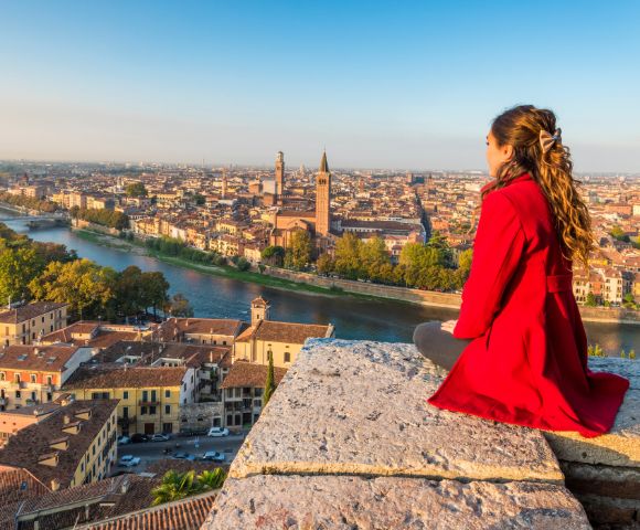Young woman in a red coat sits on a stone ledge overlooking a scenic cityscape with a river, historic buildings, and distant towers under a clear blue sky.