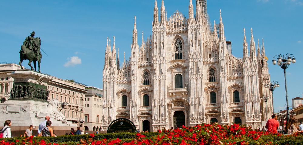 Gothic cathedral with intricate spires stands under a clear blue sky. Red flowers in the foreground and a statue to the left add contrast and depth.