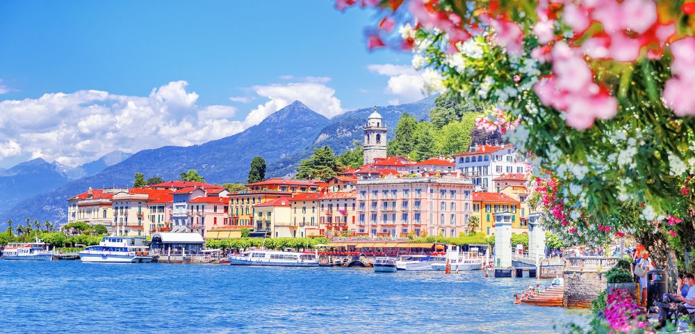 A picturesque lakeside town with colorful buildings, boats along the water, and a mountainous backdrop. Bright flowers frame the scene, evoking a vibrant, peaceful atmosphere.