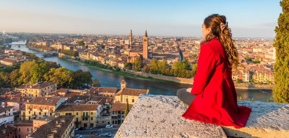 Young woman in a red coat sits on a stone ledge overlooking a scenic cityscape with a river, historic buildings, and distant towers under a clear blue sky.