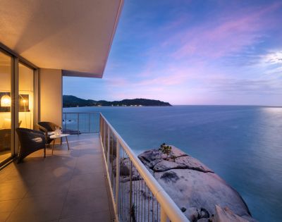 Balcony overlooking a serene ocean at sunset, with soft purple and pink hues in the sky. Two chairs and table suggest a peaceful setting.