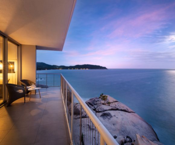 Balcony overlooking a serene ocean at sunset, with soft purple and pink hues in the sky. Two chairs and table suggest a peaceful setting.