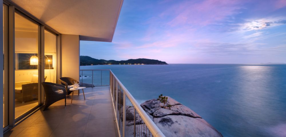 Balcony overlooking a serene ocean at sunset, with soft purple and pink hues in the sky. Two chairs and table suggest a peaceful setting.