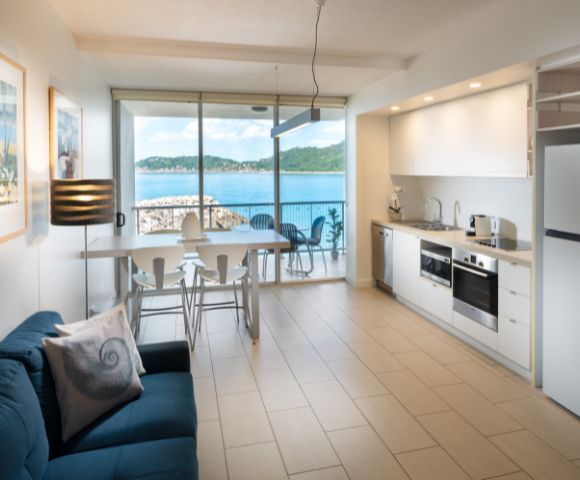 Modern apartment interior with a cozy blue sofa, a minimal white kitchen, and a dining table. Large windows reveal a stunning ocean view. Bright and serene.