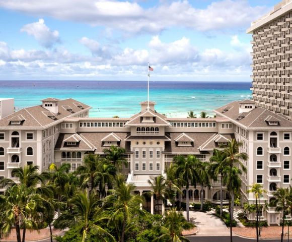 Historic beachfront hotel with a grand facade and lush palm trees, set against a vibrant blue ocean and a partly cloudy sky, conveying elegance and tranquility.