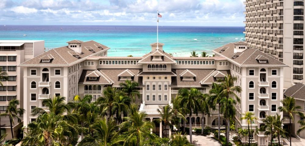 Historic beachfront hotel with a grand facade and lush palm trees, set against a vibrant blue ocean and a partly cloudy sky, conveying elegance and tranquility.