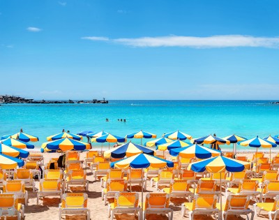 Vibrant beach scene with rows of yellow sun loungers under blue and yellow umbrellas, set against a turquoise sea and clear blue sky.