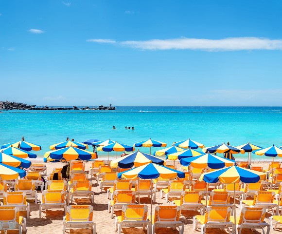Vibrant beach scene with rows of yellow sun loungers under blue and yellow umbrellas, set against a turquoise sea and clear blue sky.