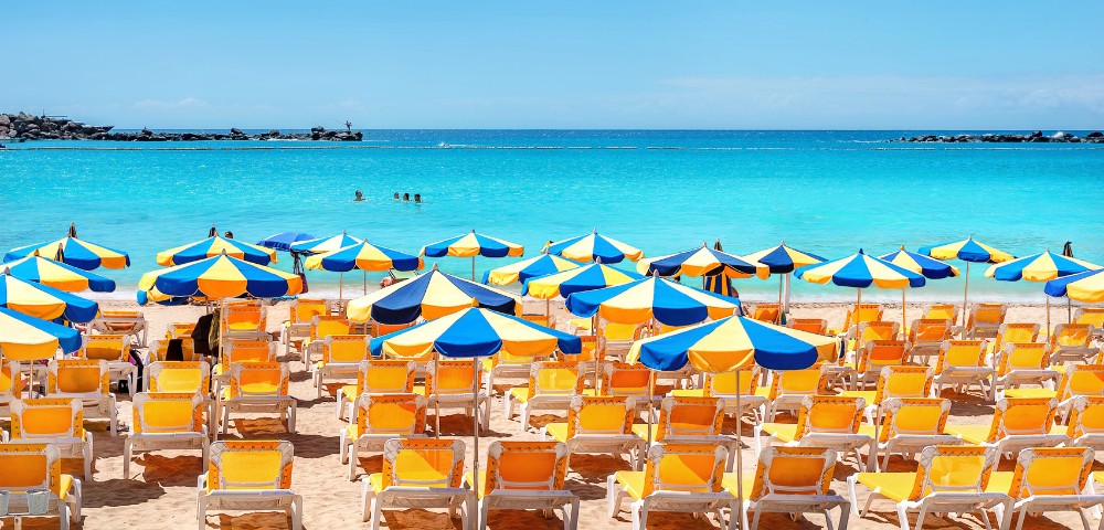 Vibrant beach scene with rows of yellow sun loungers under blue and yellow umbrellas, set against a turquoise sea and clear blue sky.