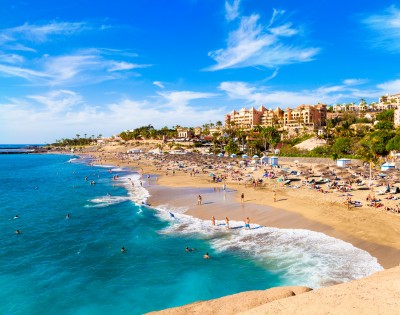 Sunny beach scene with turquoise waters and golden sand. People are swimming and sunbathing near umbrellas. Hotels and palm trees line the horizon.