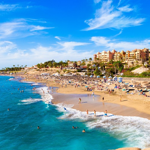 Sunny beach scene with turquoise waters and golden sand. People are swimming and sunbathing near umbrellas. Hotels and palm trees line the horizon.
