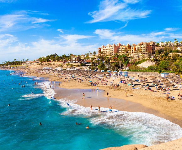 Sunny beach scene with turquoise waters and golden sand. People are swimming and sunbathing near umbrellas. Hotels and palm trees line the horizon.