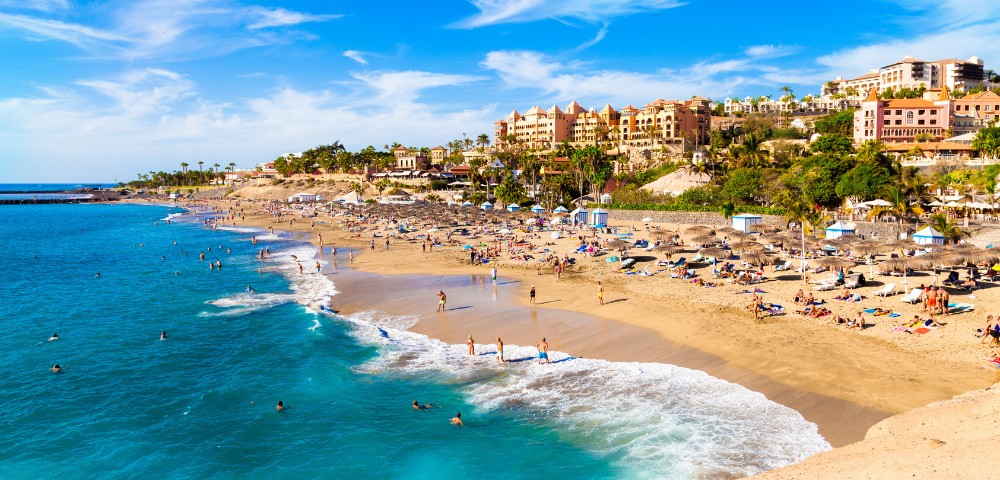 Sunny beach scene with turquoise waters and golden sand. People are swimming and sunbathing near umbrellas. Hotels and palm trees line the horizon.