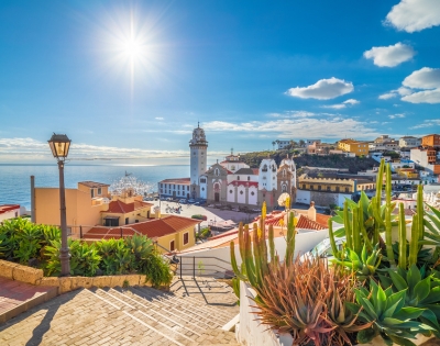 Bright coastal scene with sunny blue sky over a vibrant town. A church with a tall bell tower stands amid colorful buildings. Cacti and a lamppost in foreground.