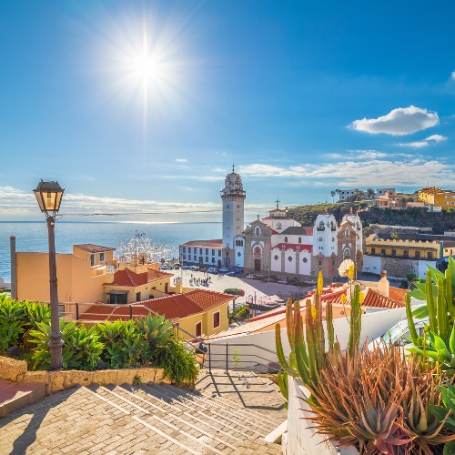 Bright coastal scene with sunny blue sky over a vibrant town. A church with a tall bell tower stands amid colorful buildings. Cacti and a lamppost in foreground.