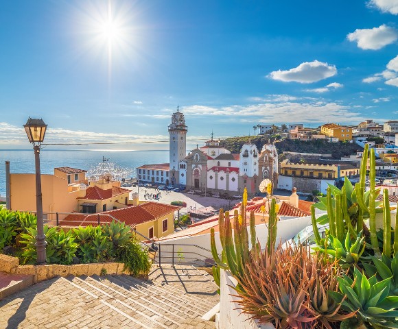 Bright coastal scene with sunny blue sky over a vibrant town. A church with a tall bell tower stands amid colorful buildings. Cacti and a lamppost in foreground.