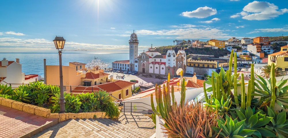 Bright coastal scene with sunny blue sky over a vibrant town. A church with a tall bell tower stands amid colorful buildings. Cacti and a lamppost in foreground.