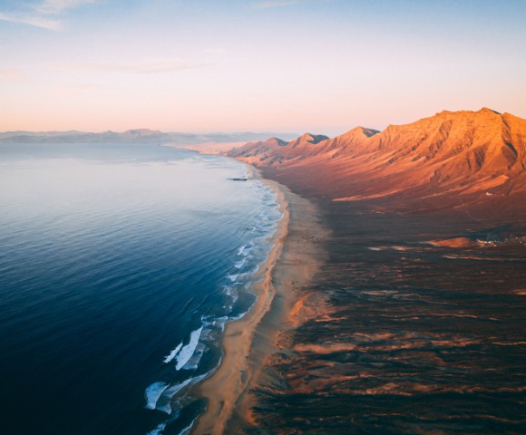 Aerial view of a vast coastline at sunrise, with gentle waves lapping a sandy beach and rugged mountains bathed in warm hues, creating a serene, expansive scene.
