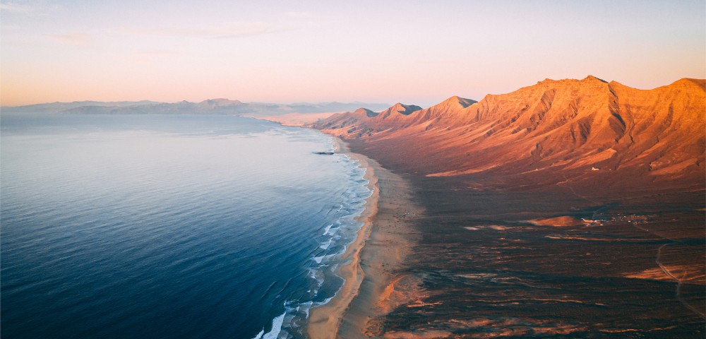 Aerial view of a vast coastline at sunrise, with gentle waves lapping a sandy beach and rugged mountains bathed in warm hues, creating a serene, expansive scene.