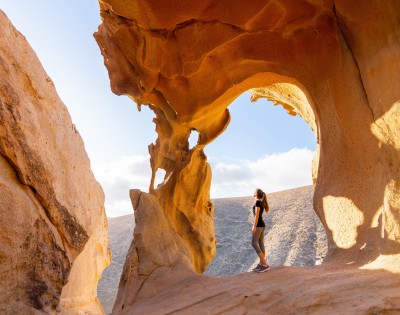 A woman stands in awe, gazing at a natural rock arch under the sun. The arch frames a clear sky, evoking a sense of wonder and tranquility.