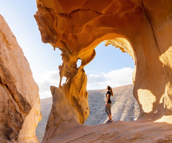 A woman stands in awe, gazing at a natural rock arch under the sun. The arch frames a clear sky, evoking a sense of wonder and tranquility.