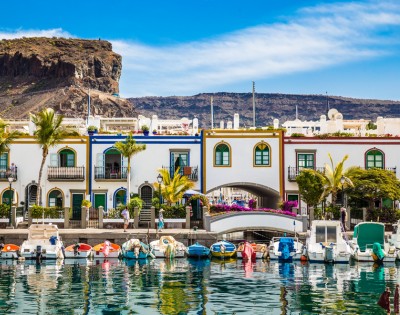 Colorful seaside village with boats in a marina, white buildings with vibrant trim, palm trees, and a rocky hill backdrop under a clear blue sky.