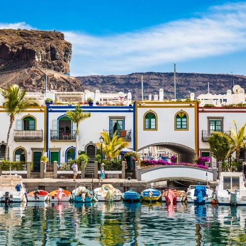 Colorful seaside village with boats in a marina, white buildings with vibrant trim, palm trees, and a rocky hill backdrop under a clear blue sky.