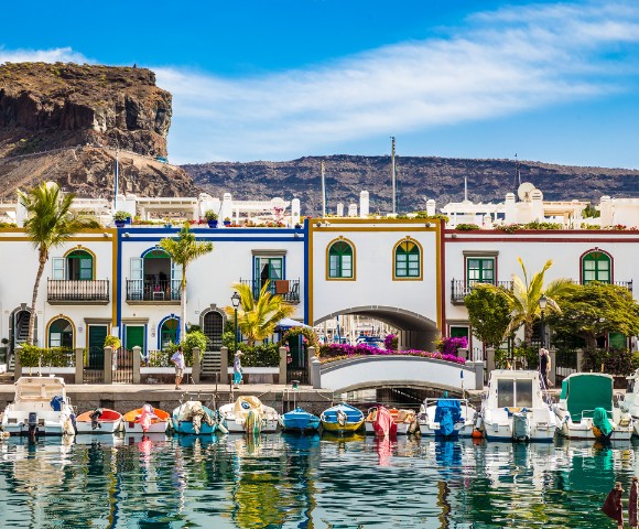 Colorful seaside village with boats in a marina, white buildings with vibrant trim, palm trees, and a rocky hill backdrop under a clear blue sky.