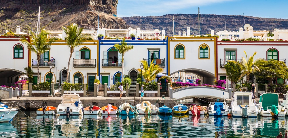 Colorful seaside village with boats in a marina, white buildings with vibrant trim, palm trees, and a rocky hill backdrop under a clear blue sky.