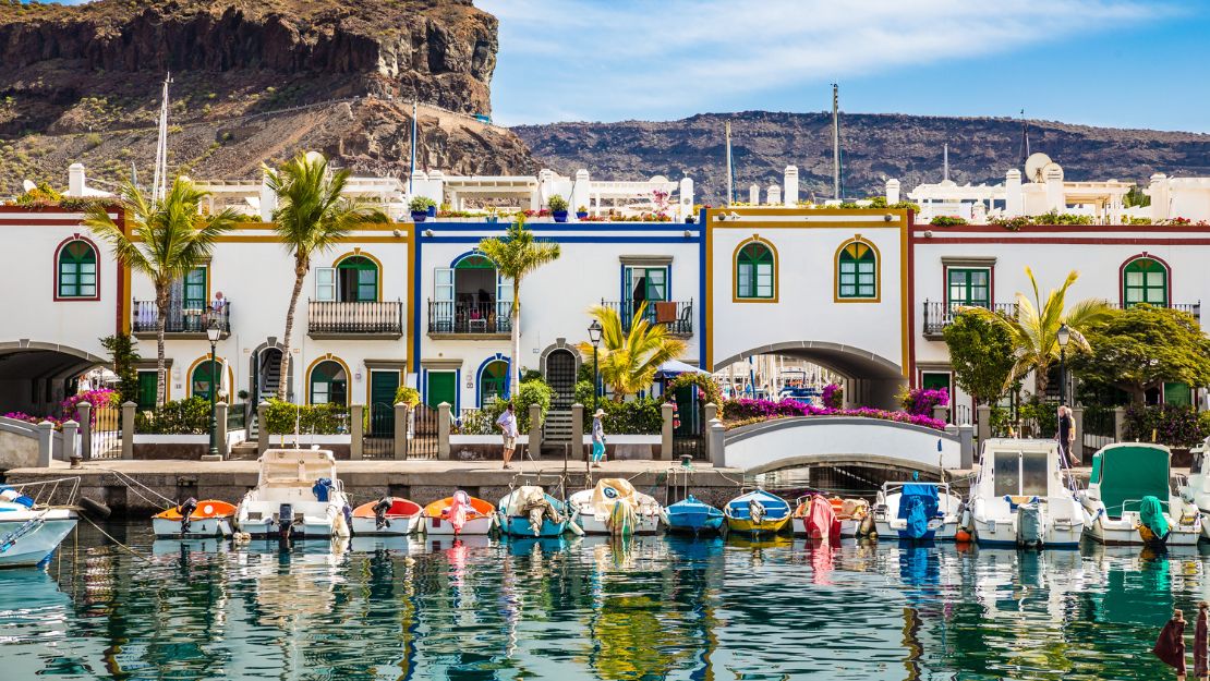 Colorful seaside village with boats in a marina, white buildings with vibrant trim, palm trees, and a rocky hill backdrop under a clear blue sky.