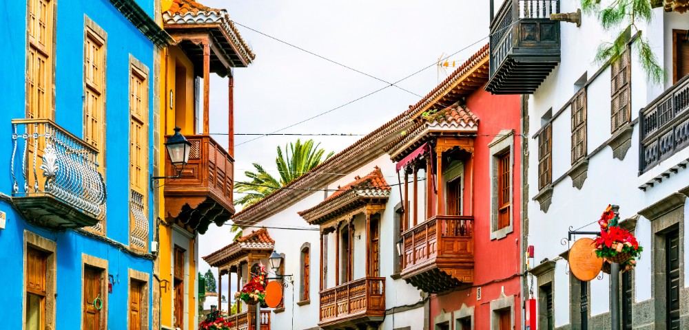 Colorful street with vibrant blue, red, and white buildings featuring wooden balconies. Hanging flower baskets add charm, creating a lively, welcoming atmosphere.