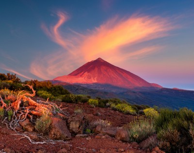 Vibrant sunset over a volcanic mountain, with swirling clouds above. Red-orange light casts a warm glow on rocky terrain and sparse vegetation in the foreground.