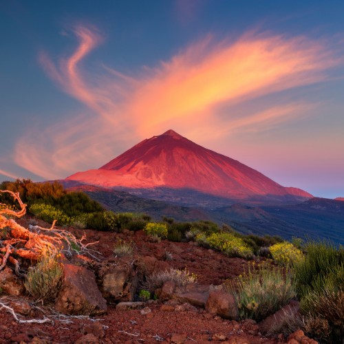 Vibrant sunset over a volcanic mountain, with swirling clouds above. Red-orange light casts a warm glow on rocky terrain and sparse vegetation in the foreground.