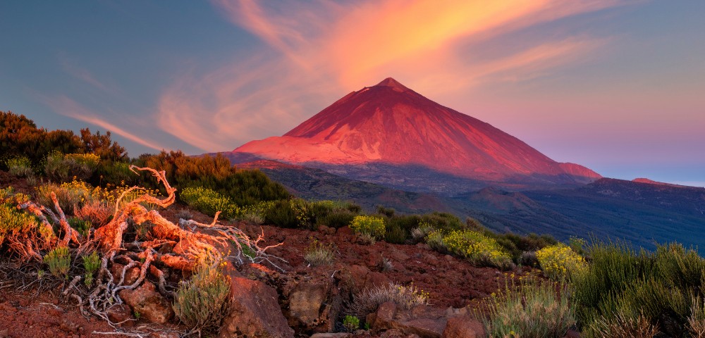 Vibrant sunset over a volcanic mountain, with swirling clouds above. Red-orange light casts a warm glow on rocky terrain and sparse vegetation in the foreground.