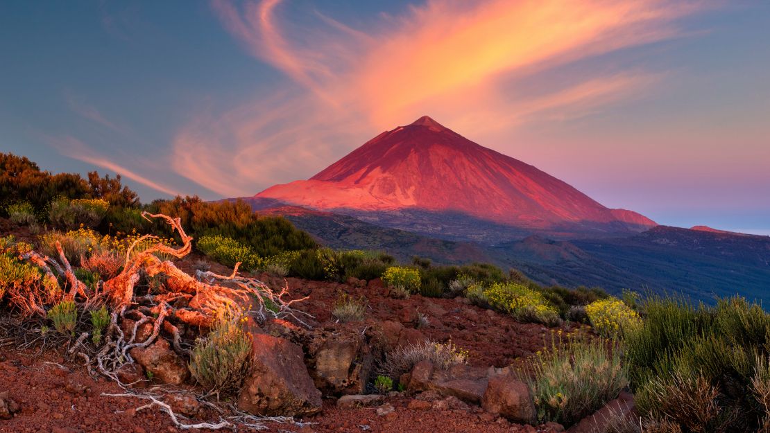 Vibrant sunset over a volcanic mountain, with swirling clouds above. Red-orange light casts a warm glow on rocky terrain and sparse vegetation in the foreground.