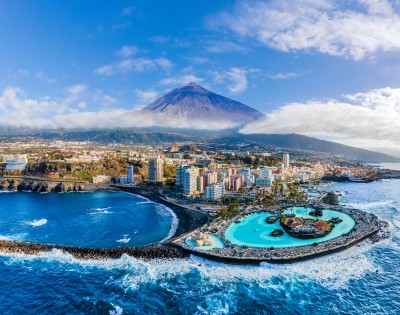 Aerial view of a coastal city with a large pool adjacent to the ocean. Tall buildings are nestled at the base of a mountain under a clear blue sky.