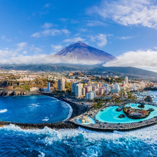 Aerial view of a coastal city with a large pool adjacent to the ocean. Tall buildings are nestled at the base of a mountain under a clear blue sky.