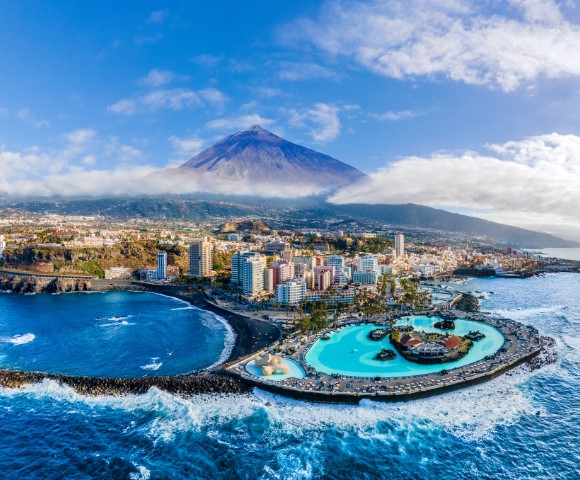 Aerial view of a coastal city with a large pool adjacent to the ocean. Tall buildings are nestled at the base of a mountain under a clear blue sky.