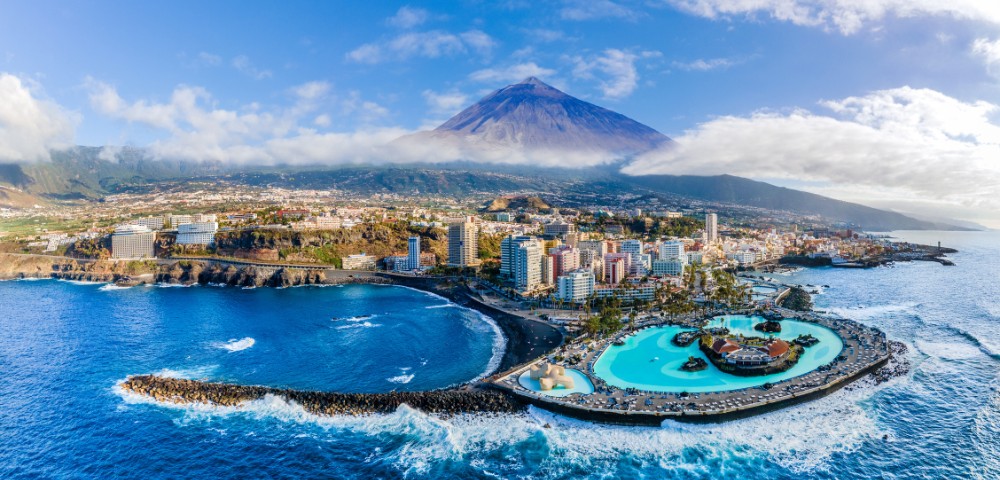 Aerial view of a coastal city with a large pool adjacent to the ocean. Tall buildings are nestled at the base of a mountain under a clear blue sky.
