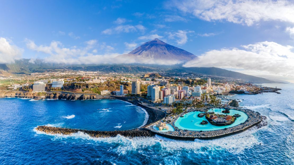 Aerial view of a coastal city with a large pool adjacent to the ocean. Tall buildings are nestled at the base of a mountain under a clear blue sky.